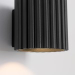 Kinkiet Aura 1xG9 w kolorze czarnym, z cylindrycznym abażurem o żłobionej fakturze. Widoczny metalowy uchwyt montażowy. Oświetlenie skierowane w dół, na jasnym tle.