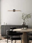 Kinkiet Calaca 1xLED 8W w kolorze czarnym, zamontowany na ścianie. Posiada prostokątną, metalową obudowę z delikatnym, złotym akcentem. W tle widoczna aranżacja wnętrza z meblami w odcieniach czerni i naturalnego drewna.