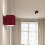 Kinkiet Aura 1xG9 w kolorze burgundowym, o falistej fakturze, zamontowany na ścianie. Oświetlenie emitowane jest z dolnej części lampy. W tle widoczne zasłony oraz okno, co podkreśla domowy klimat.