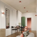 Lampa sufitowa Aura w kolorze brzoskwiniowym, zamontowana na suficie. Posiada cylindryczny kształt i jest umieszczona w nowoczesnym wnętrzu z drewnianą podłogą, stołem i krzesłami. W tle znajduje się zielona szafka oraz lustro.