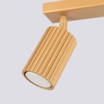 Lampa sufitowa Karbon w kolorze złotym, z dwoma reflektorami GU10. Ujęcie z boku na detal cylindrycznego klosza z rowkowaną fakturą. Montaż na suficie, minimalistyczny design. Tło w jasnym odcieniu.