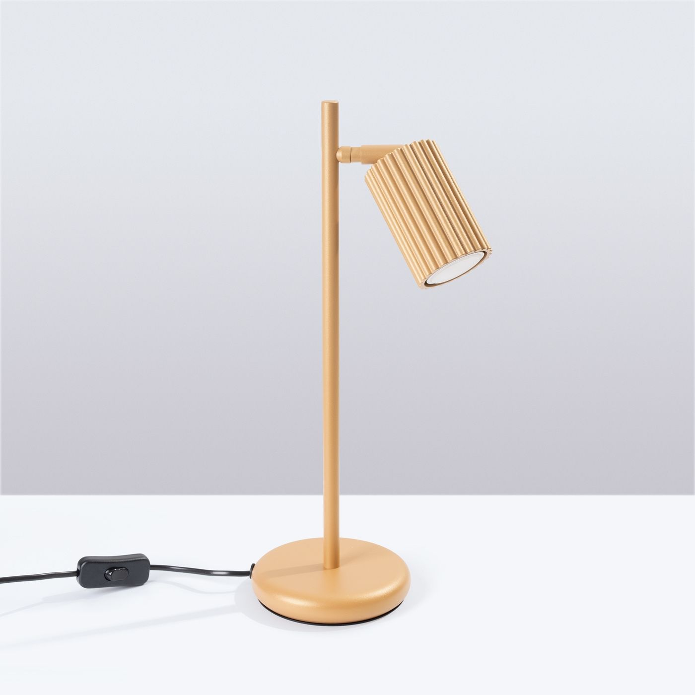 Lampka stołowa Karbon 1xGU10 w kolorze złotym, z cylindryczną, żebrowaną głowicą, zamontowana na prostym, okrągłym stojaku. Widoczny kabel z włącznikiem w kolorze czarnym. Produkt ustawiony na białym tle.