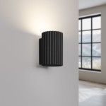 Kinkiet Karbon 10 w kolorze czarnym, z cylindryczną, żebrowaną obudową, montowany na ścianie. Źródło światła GU10, skierowane ku górze, w nowoczesnym wnętrzu z oknem w tle.