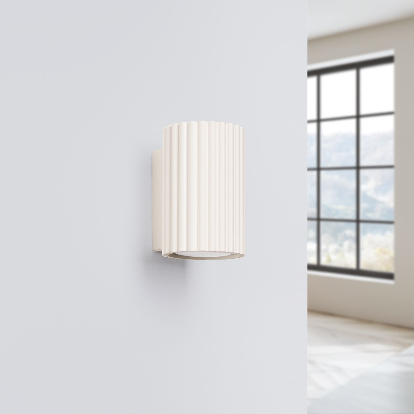 Beżowy kinkiet Karbon 10 z oprawą w kształcie cylindrycznego klosza z pionowymi żłobieniami, montowany na ścianie. Widoczny pojedynczy punkt świetlny GU10. Tło w jasnym wnętrzu z dużym oknem.