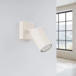 Kinkiet Ring 1xGU10 w kolorze beżowym, zamontowany na ścianie. Okrągła głowica lampy skierowana w dół, prostokątna baza. Tło jasne, z oknem w oddali, co podkreśla nowoczesny design.