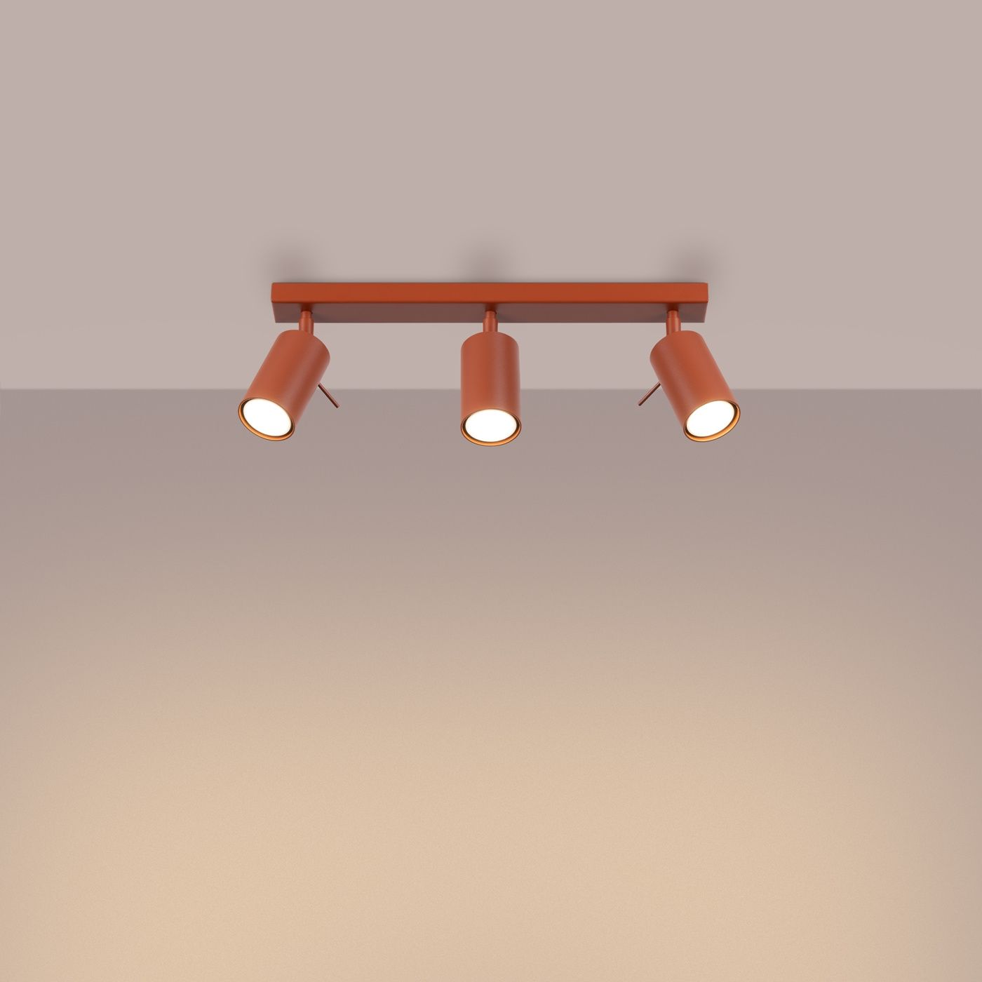 Lampa sufitowa Ring w kolorze ochra czerwona z trzema reflektorami GU10, zamontowana na prostokątnej podstawie. Reflektory mają cylindryczny kształt i są skierowane w dół, tworząc równomierne oświetlenie. Tło w jasnym odcieniu.