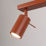 Lampa sufitowa spot w kolorze ochra czerwona, z metalowym kloszem w kształcie cylindra. Ujęcie z bliska, pokazujące detal lampy z widocznym uchwytem. Element montażowy w prostokątnej podstawie.