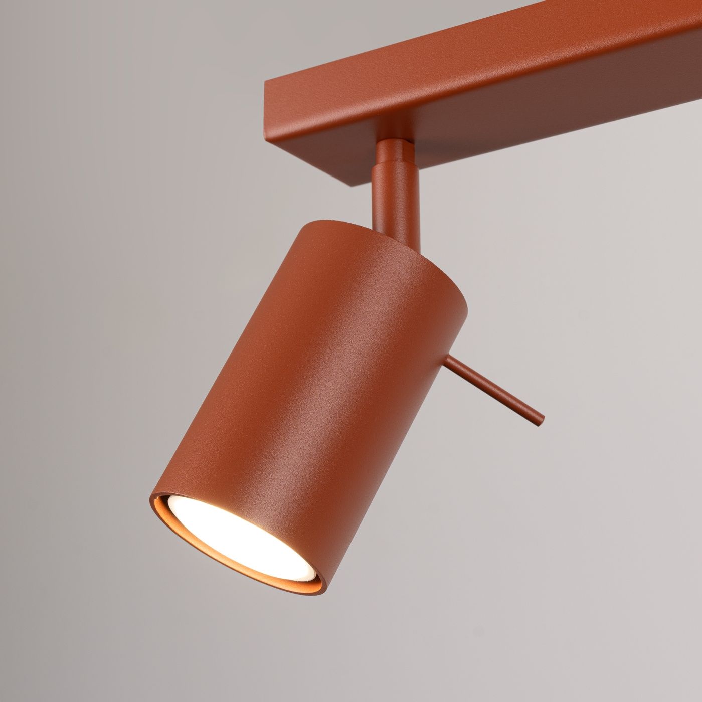 Lampa sufitowa typu spot w kolorze ochra czerwona, z metalowym wykończeniem. W kształcie cylindrycznym, z widocznym źródłem światła GU10. Ujęcie zbliżeniowe, ukazujące detale konstrukcji i teksturę powierzchni.