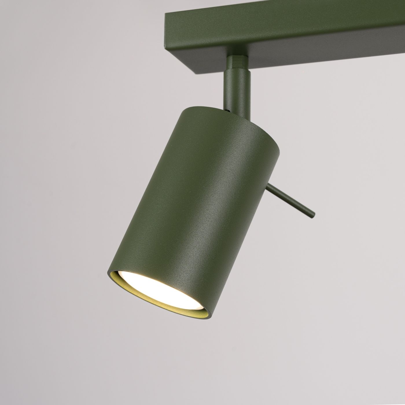 Lampa sufitowa typu spot w kolorze oliwkowym, z metalowym kloszem w formie cylindra. Ujęcie zbliżeniowe, ukazujące detal klosza z widocznym źródłem światła. Element montażowy w prostokątnej podstawie.