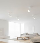 Biała lampa sufitowa w stylu spot, jednolita konstrukcja z okrągłym kloszem, zamontowana na prostokątnej podstawie. Ujęcie z dołu, ukazujące jej minimalistyczny design w jasnym wnętrzu z oknami. Idealna do oświetlenia przestrzeni.