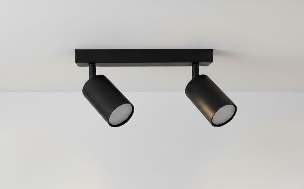 Czarna lampa sufitowa (spot) 2-punktowa z metalowymi kloszami, zamontowana na prostokątnej podstawie. Klosze są cylindryczne, skierowane w różnych kierunkach, co umożliwia regulację oświetlenia. Tło jasne, jednolite.
