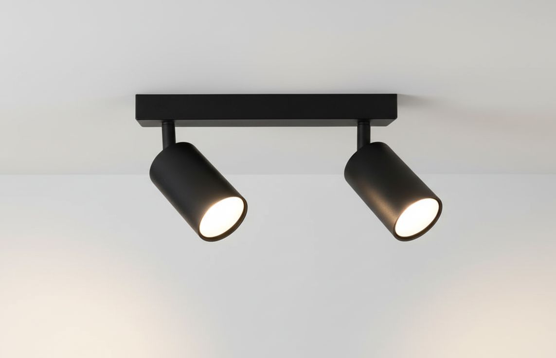 Czarna lampa sufitowa typu spot z dwoma ruchomymi głowicami, zamontowana na prostokątnej podstawie. Głowice mają cylindryczny kształt i emitują jasne światło. Ujęcie z góry, na jednolitym tle.