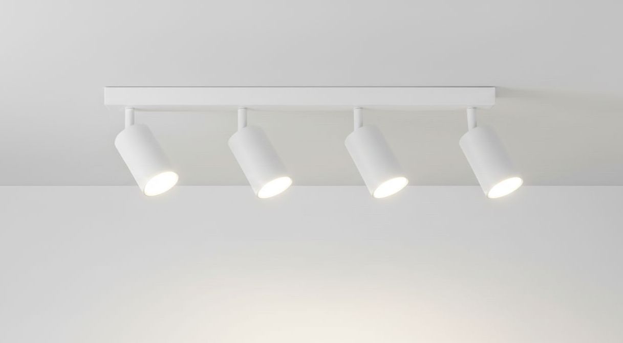 Biała lampa sufitowa 4-punktowa z czterema ruchomymi reflektorami na prostokątnej podstawie. Reflektory mają cylindryczny kształt, skierowane w dół, montowane do sufitu. Tło proste, jasne.