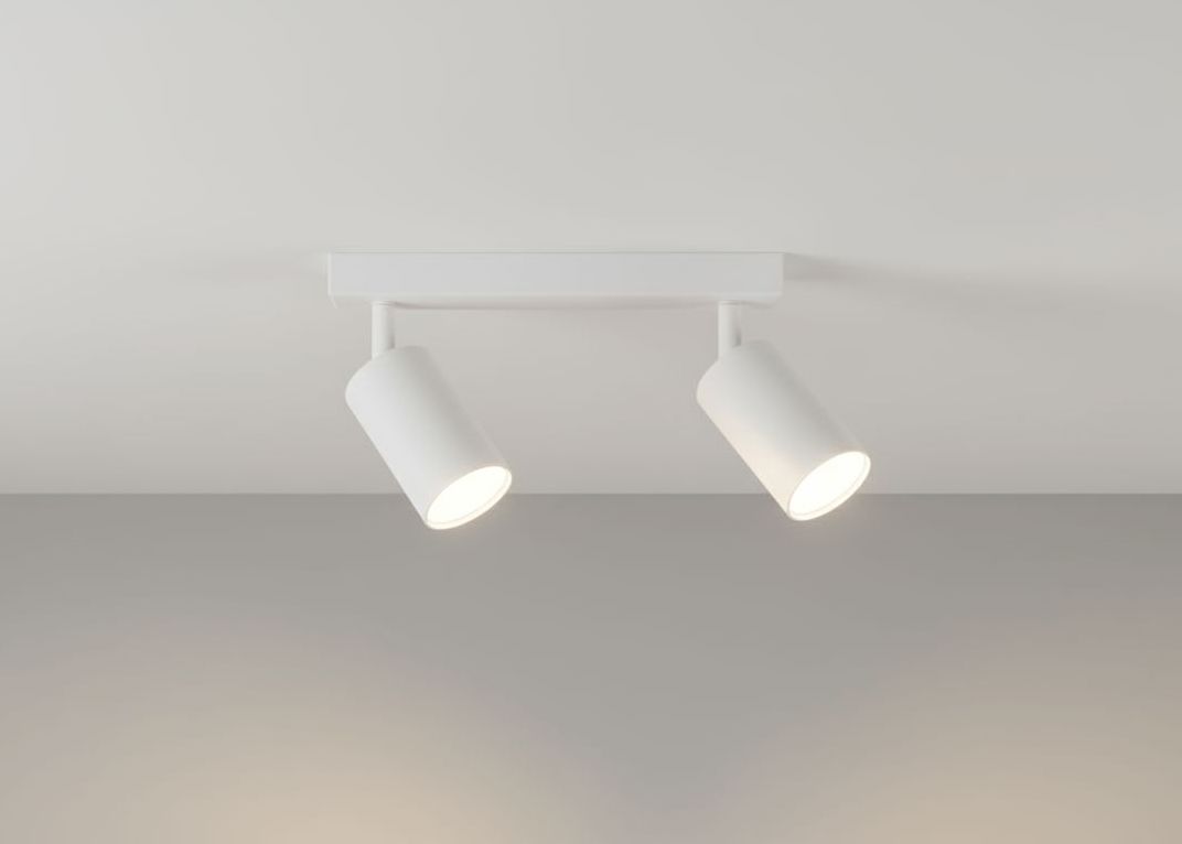 Biała lampa sufitowa typu spot z dwoma punktami świetlnymi, zamontowana na prostokątnej podstawie. Ujęcie z dołu, oba reflektory skierowane w przeciwnych kierunkach, minimalistyczny design. Tło w neutralnym, jasnym kolorze.