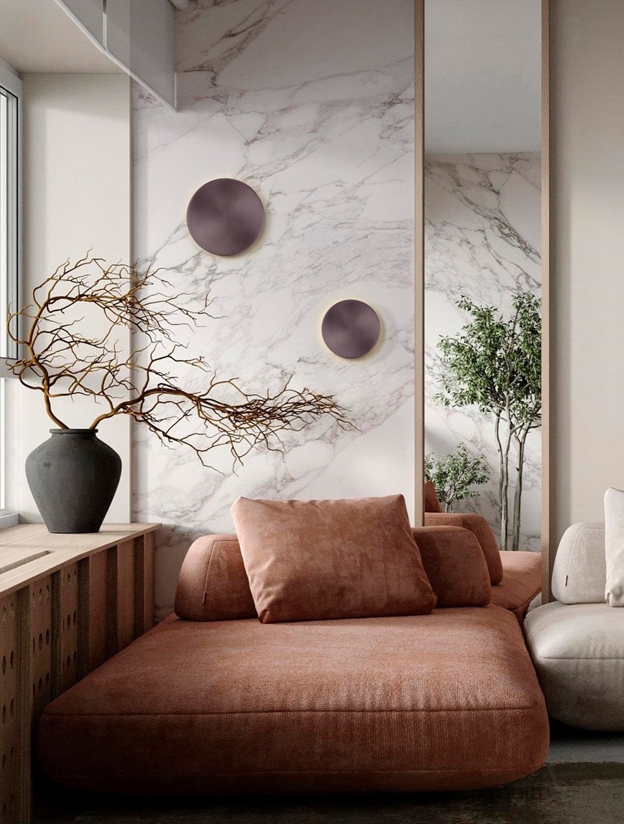 Kinkiet Chica w kolorze brązowym, z pojedynczym źródłem światła LED o mocy 21W. Produkt zamontowany na ścianie, z minimalistycznym designem, idealny do nowoczesnych wnętrz. Tło w jasnych odcieniach z elementami roślinnymi i dekoracyjnymi.
