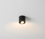 Mini lampa sufitowa tuba w kolorze czarnym, zamontowana na białym suficie. Okrągły kształt z widocznym źródłem światła, skierowany w dół, emitujący ciepłe światło. Prosty design, idealna do oświetlenia wnętrz.