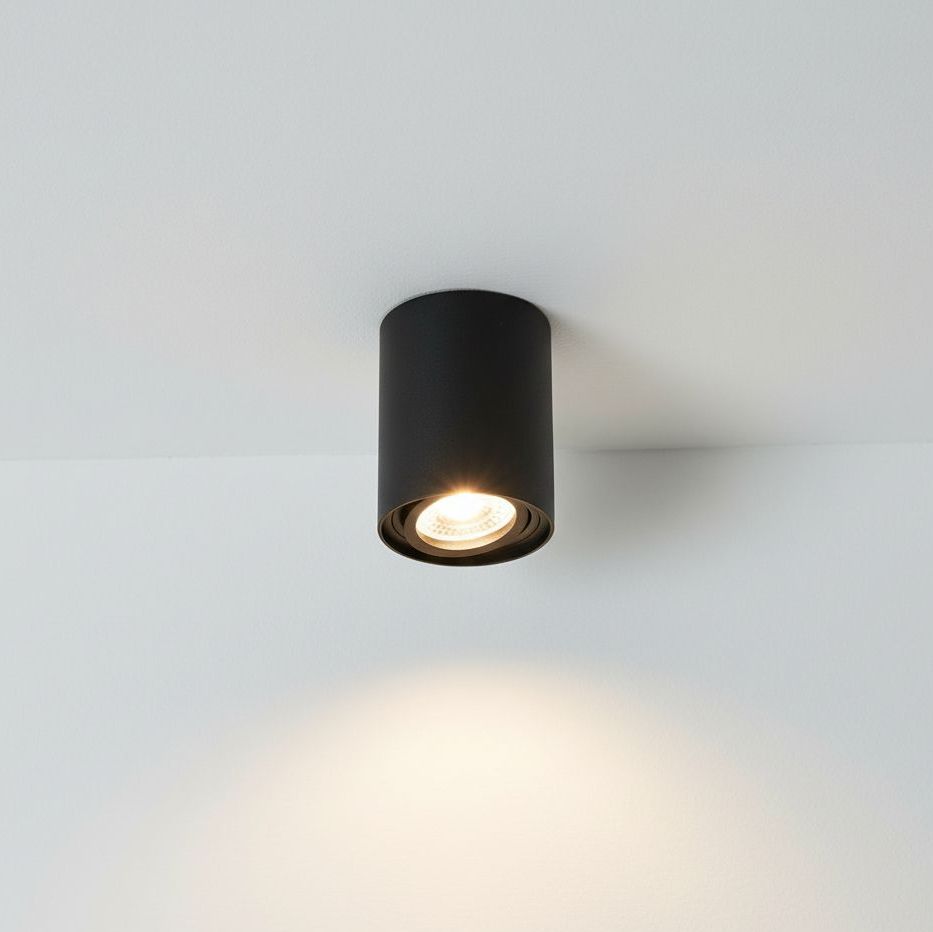 Czarna lampa sufitowa w kształcie tuby, montowana na suficie. Oświetlenie skierowane jest w dół, podkreślając prosty, minimalistyczny design. Metaliczna faktura obudowy nadaje jej nowoczesny wygląd. Tło jasne, jednolite.