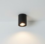 Czarna lampa sufitowa w kształcie tuby, montowana na suficie. Oświetlenie skierowane jest w dół, podkreślając prosty, minimalistyczny design. Metaliczna faktura obudowy nadaje jej nowoczesny wygląd. Tło jasne, jednolite.