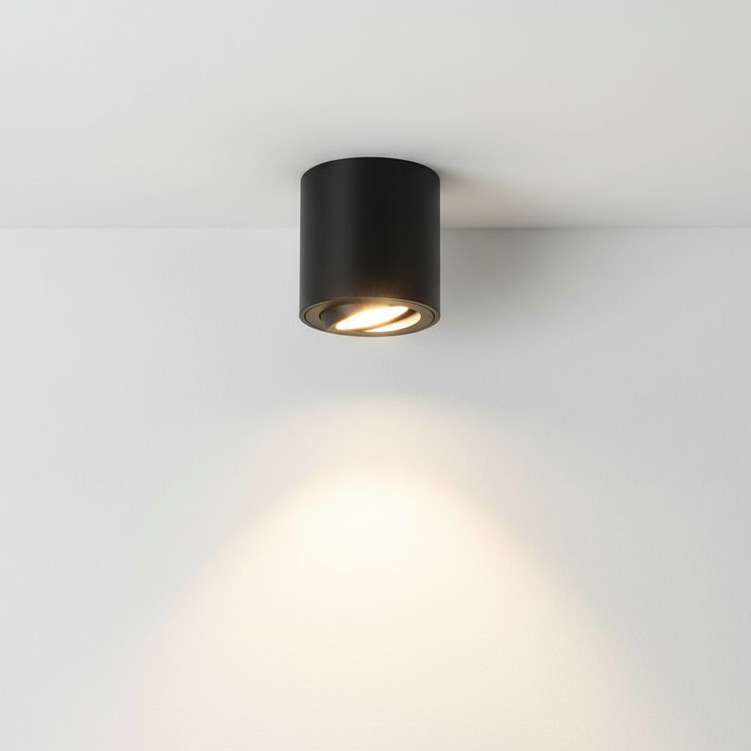 Czarna lampa sufitowa w formie tuby, zamontowana na białym suficie, z widocznym źródłem światła skierowanym w dół. Emituje jasne światło, tworząc delikatny cień na ścianie. Prosta, cylindryczna konstrukcja, idealna do nowoczesnych wnętrz.