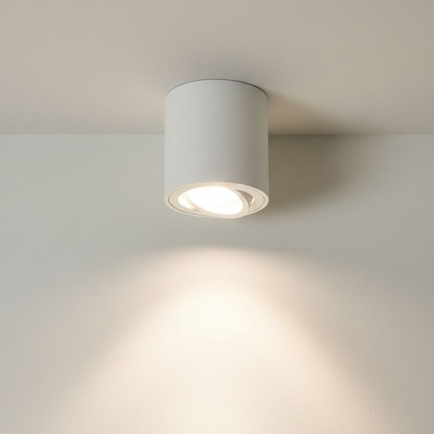 Biała lampa sufitowa typu tuba w formie cylindrycznej, zamontowana na suficie, z widocznym źródłem światła w środku. Emituje jasne światło skierowane w dół, na tle gładkiej, jasnej ściany.