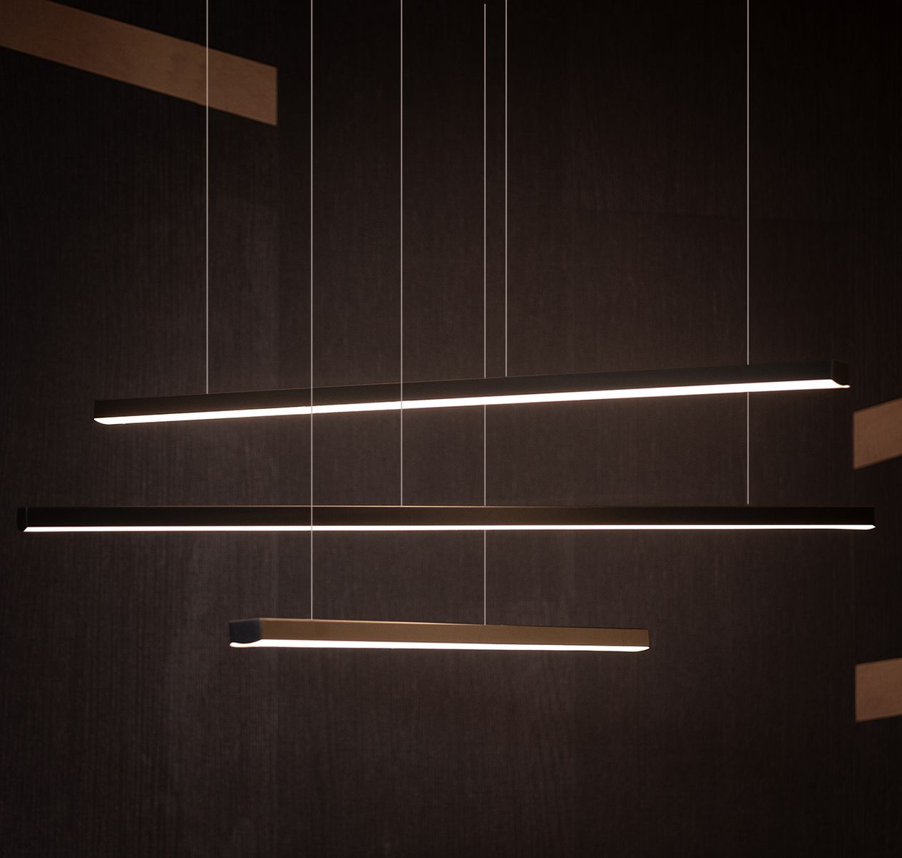 Trio LED lampa wisząca ściemnialna, czarna, składająca się z trzech poziomych prostokątnych elementów. Zawieszona na cienkich linkach, z ciepłym światłem LED emitowanym z górnej krawędzi. Tło ciemne, minimalistyczne.
