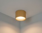 Lampa sufitowa Gisele w złotym kolorze, w kształcie tuby, z teksturowaną powierzchnią. Ujęcie z dołu, zamontowana w rogu pomieszczenia, emitująca ciepłe światło. Proste, nowoczesne wzornictwo, idealna do wnętrz.