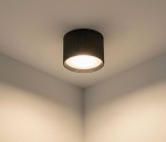 Czarna lampa sufitowa tuba GX53, zamontowana w kącie sufitu. Okrągła, teksturowana obudowa z widocznym źródłem światła. Prosta, nowoczesna forma, na jasnym tle ściany.