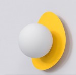 Kinkiet Ovo w kolorze pomarańczowym, z białą kulistą lampą i asymetrycznym, żółtym tłem. Produkt zamontowany na gładkiej, jasnej ścianie, widoczny z przodu. Okrągły kształt lampy oraz kontur tła tworzą nowoczesny design.