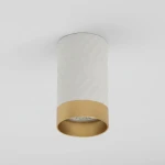 Lampa sufitowa Arras w kształcie tuby, z białym, teksturowanym korpusem i złotym wykończeniem u dołu. Oświetlenie z miejscem na jedną żarówkę GU10, montowane na suficie, widoczne zbliżenie na detal klosza i oprawy. Tło w jasnym odcieniu.