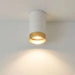 Lampa sufitowa Arras w formie tuby, z białym korpusem w wzór chevron i złotym wykończeniem u dołu. Źródło światła GU10, montowana na suficie, emituje ciepłe światło, tworząc przyjemną atmosferę. Tło jasne, minimalistyczne.