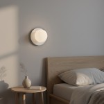 Biały kinkiet Americana 1xG9, okrągły, zamontowany na ścianie w sypialni. W tle widoczna drewniana łóżko z poduszkami i stolik nocny z wazonem oraz książką. Delikatne światło rzucane na ścianę.