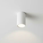 Boka lampa sufitowa w kształcie białej tuby, zamontowana na suficie, z miejscem na jedną żarówkę GU10. Widoczna jasna oprawa emitująca światło, z gładką, matową powierzchnią.