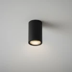 Czarna lampa sufitowa w kształcie tuby, zamontowana na białym suficie. Posiada jedno gniazdo GU10, emitujące ciepłe światło. Prosta, cylindryczna forma z matowym wykończeniem, idealna do nowoczesnych wnętrz.