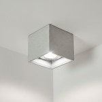 Lampa sufitowa Bold M w kształcie sześcianu, wykonana z betonu w jasnym odcieniu, z okrągłym źródłem światła wewnątrz. Ujęcie z dołu, ukazujące montaż na suficie. Prosta, nowoczesna forma, idealna do minimalistycznych wnętrz.