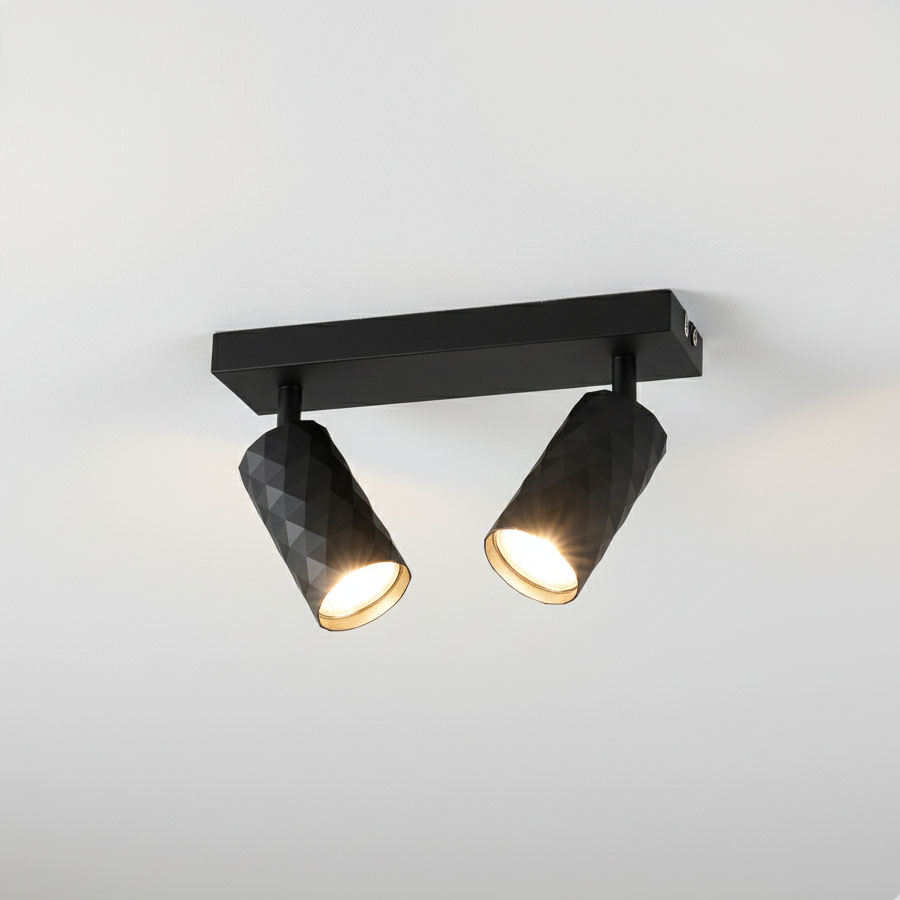 Czarna lampa sufitowa typu spot z dwoma reflektorami GU10 o geometrycznym, teksturowanym kształcie. Ujęcie z dołu, zamontowana na ścianie, z widocznym światłem emitowanym przez obie lampy. Prosta, nowoczesna konstrukcja na jednolitym tle.