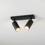 Czarna lampa sufitowa typu spot z dwoma reflektorami GU10 o geometrycznym, teksturowanym kształcie. Ujęcie z dołu, zamontowana na ścianie, z widocznym światłem emitowanym przez obie lampy. Prosta, nowoczesna konstrukcja na jednolitym tle.
