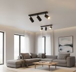 Lampa sufitowa spot Glamour 3xGU10 w kolorze czarnym, zamontowana na suficie. Posiada trzy ruchome reflektory, które oświetlają przestrzeń. W tle widoczny nowoczesny salon z szaro-beżową sofą i dużymi oknami.