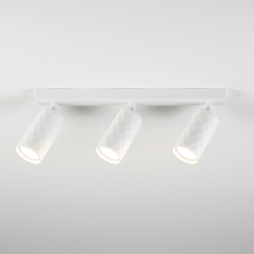 Biała lampa sufitowa typu spot z trzema reflektorami GU10, zamocowana na prostokątnej podstawie. Reflektory mają diamentową fakturę, skierowane w różnych kierunkach, co pozwala na regulację oświetlenia. Tło jest jasne i neutralne.