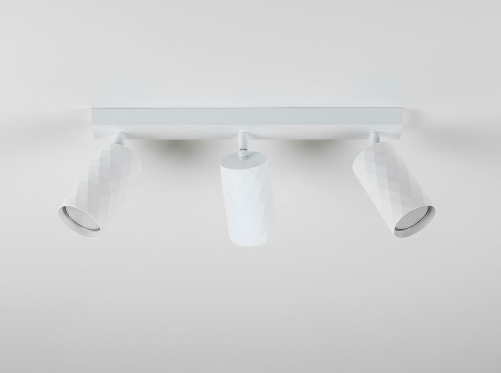 Lampa sufitowa spot z trzema reflektorami w kolorze białym, wykonana z materiału o fakturze diamentowej. Reflektory zamocowane na prostokątnym stelażu, skierowane w różnych kierunkach. Ujęcie z przodu, na jasnym tle.