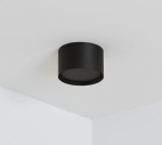 Lampa sufitowa Cyklop w kształcie czarnej tuby, zamontowana na suficie. Posiada miejsce na jedną żarówkę GX53. Prosty, okrągły kształt i matowe wykończenie. Tło jasne, minimalistyczne.