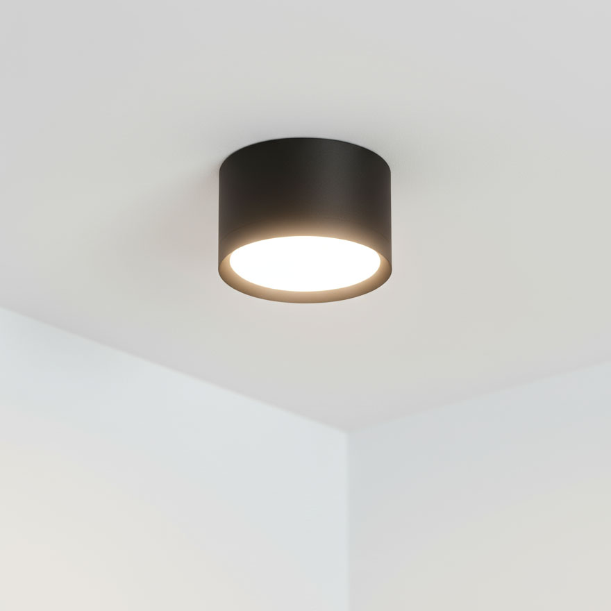 Lampa sufitowa Cyklop w kształcie czarnej tuby, z jedną oprawą GX53. Zainstalowana pod sufitem, emitująca jasne światło, z minimalistycznym designem pasującym do nowoczesnych wnętrz. Tło jasne, neutralne.
