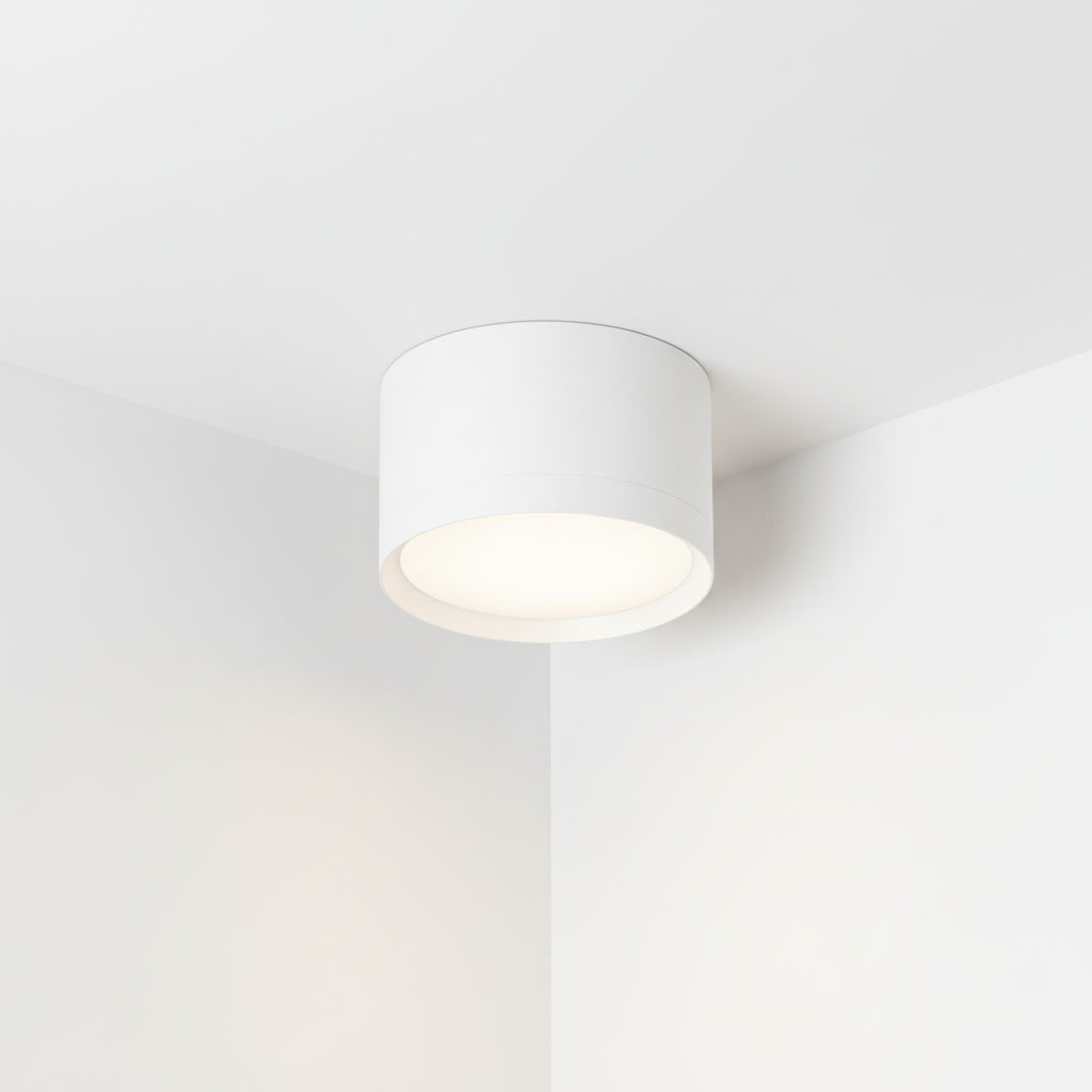 Lampa sufitowa Cyklop w kształcie tuby, kolor biały, z miejscem na jedną żarówkę GX53. Ujęcie z góry, zamontowana w rogu pomieszczenia. Prosty design, gładka powierzchnia, matowe wykończenie, emitująca jasne światło.