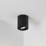 Lampa sufitowa tuba kierunkowa Devo w kolorze czarnym, o cylindrycznym kształcie, zamontowana na suficie w rogu pomieszczenia. Wnętrze lampy zawiera oprawkę na żarówkę GU10, skierowaną w dół. Tło jest jasne i jednolite.