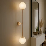 Kinkiet Lumia 2xE27 w kolorze złotym i białym, z dwoma kulistymi kloszami z mlecznego szkła zawieszonymi na cienkiej, złotej ramie. Produkt zamontowany na ścianie, obok lustra, w nowoczesnym wnętrzu.