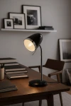 Lampka biurkowa Kajtek w kolorze czarnym z metalowym, elastycznym ramieniem i okrągłą podstawą. Widoczna żarówka E27, na drewnianym biurku w minimalistycznym wnętrzu z książkami i dekoracjami w tle.