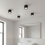 Haro lampa sufitowa tuba GX53/IP54 w kolorze czarnym, zamontowana na suficie. Okrągła forma z białym kloszem, umieszczona w minimalistycznym wnętrzu łazienki z białymi kafelkami i dużym lustrem.