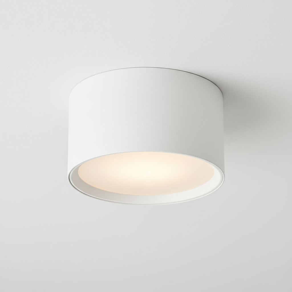 Biała lampa sufitowa w kształcie tuby, montowana na suficie. Posiada oprawę na jedną żarówkę GX53. Kąt ujęcia z góry, ukazujący okrągłą formę oraz jasne światło emitowane przez wnętrze lampy. Tło w kolorze białym.