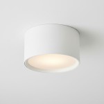 Biała lampa sufitowa w kształcie tuby, montowana na suficie. Posiada oprawę na jedną żarówkę GX53. Kąt ujęcia z góry, ukazujący okrągłą formę oraz jasne światło emitowane przez wnętrze lampy. Tło w kolorze białym.