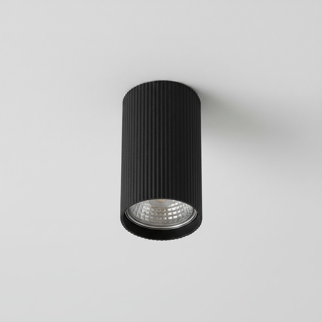Lucerno lampa sufitowa w kształcie tuby, czarna, z rowkowaną teksturą na obudowie. Umieszczona na suficie, z widoczną żarówką GU10 i reflektorem, emitującym światło w dół. Tło w jasnym kolorze, minimalistyczne.