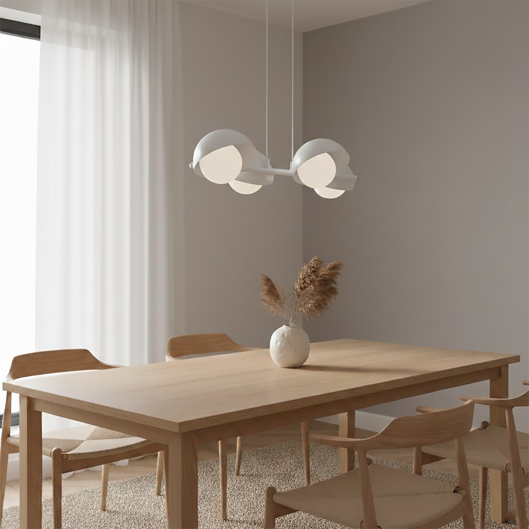 Biała lampa wisząca Boom z czterema kloszami G9 zawieszona nad drewnianym stołem w jasnym wnętrzu. Klosze mają okrągły kształt i minimalistyczny design. W tle widoczne krzesła oraz wazon z roślinami ozdobnymi.